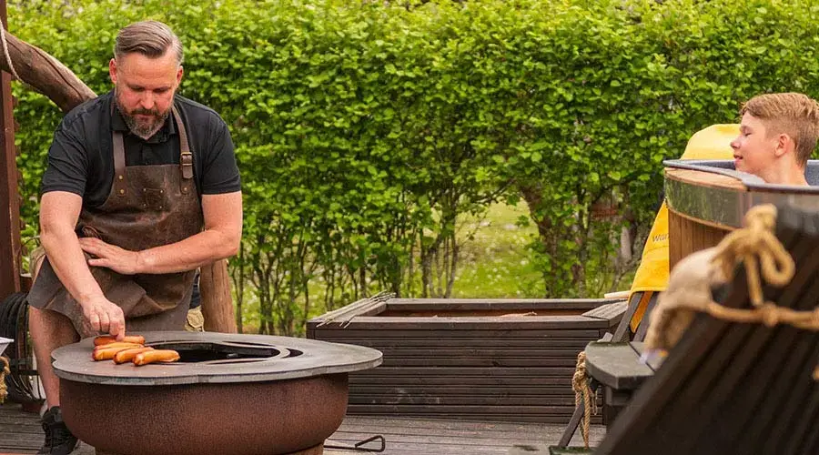 Un homme barbecue des saucisses près d'un jeune garçon, dans un jardin verdoyant.